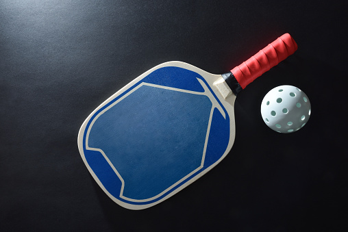 Blue and red wooden paddle pickleball racket and a white ball on black table. Top view.