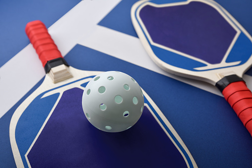 Blue and red wooden pickleball paddles and a white ball on blue playing court. Elevated view.