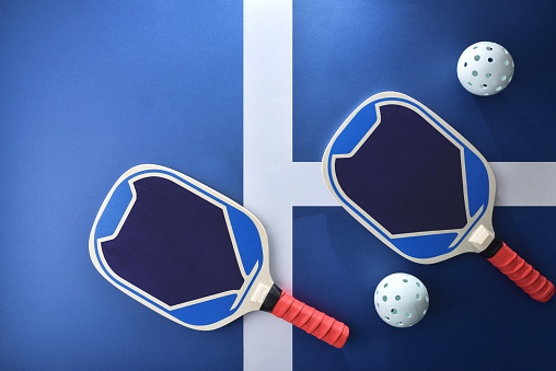 Blue and red wooden pickleball paddles and a white balls on blue playing court. Top view.