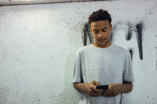 A medium close-up of a young teenage man stood in an underpass waiting for friends. He is standing looking down and using his mobile phone. The underpass is in Newcastle upon Tyne in the North East of England.