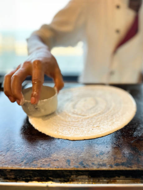 Image of unrecognisable chef in chef whites using plastic cup to spread dosa pancake batter on hot stove hotplate, commercial kitchen setting, elevated view, focus on foreground stock photo