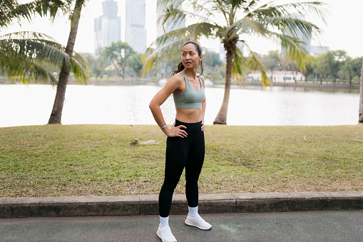 Portrait of healthy young woman in sports clothing with hands on hips standing at the park. Confident young female taking break from workout at park.
