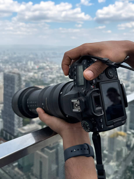istock Close-up image unrecognisable person on observation deck, taking cityscape photographs with digital SLR camera, zoom lens balanced on glass safety barrier railing, skyscraper skyline, downtown, city centre Bangkok, Thailand, sunny blue sky, fluffy clouds 2148410997