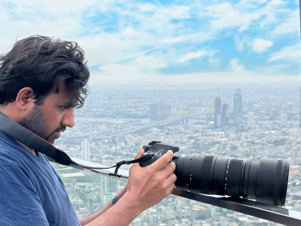 istock Close-up image man on observation deck, taking cityscape photographs with digital SLR camera, zoom lens balanced on glass safety barrier railing, skyscraper skyline view, Sukhumvit downtown, city centre Bangkok, Thailand, sunny blue sky, fluffy clouds 2148411027