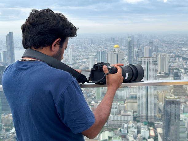 istock Close-up image man on observation deck, taking cityscape photographs with digital SLR camera, zoom lens balanced on glass safety barrier railing, skyscraper skyline view, Sukhumvit downtown, city centre Bangkok, Thailand, sunny blue sky, fluffy clouds 2148411042