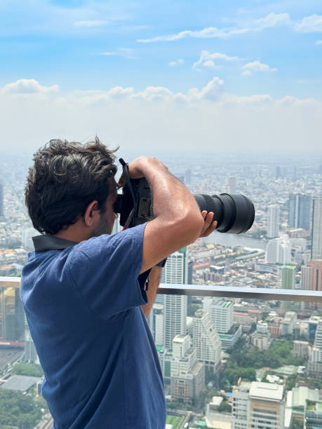 istock Image man on observation deck, glass safety barrier, taking cityscape photographs with digital SLR camera using zoom lens, skyscraper skyline view, Sukhumvit downtown, city centre Bangkok, Thailand, sunny blue sky with fluffy white clouds 2148411090