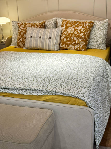 istock Close-up image of hotel room double bed with stack of floral patterned pillows leaning on headboard, grey and white folded duvet cover, yellow bed sheets, bedroom bench at foot of bed, wooden wall panelling, focus on foreground 2148411334