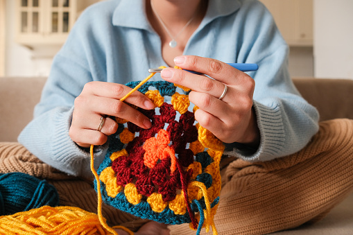 Young woman knitting crocheting with colored yarn granny square at home. Woman doing needlework, home hobbies. High quality photo
