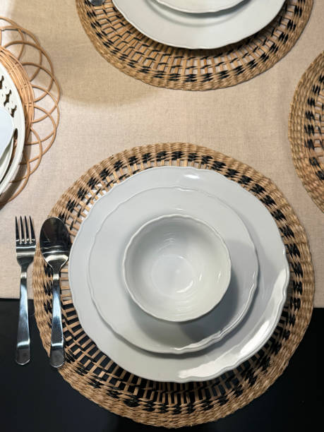 istock Close-up image of dining table set for breakfast / lunch / dinner with stacked white dinner plates, side plates and bowls on round wicker placemats, stainless steel cutlery spoons and forks, elevated view 2148414884