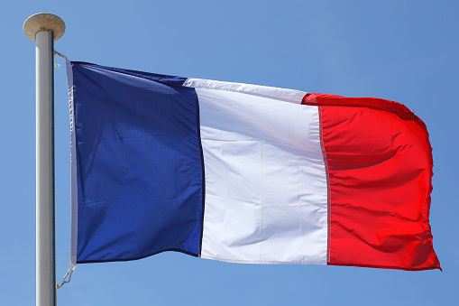 The flag of France waves on a flagpole against a blue sky on a sunny day.
