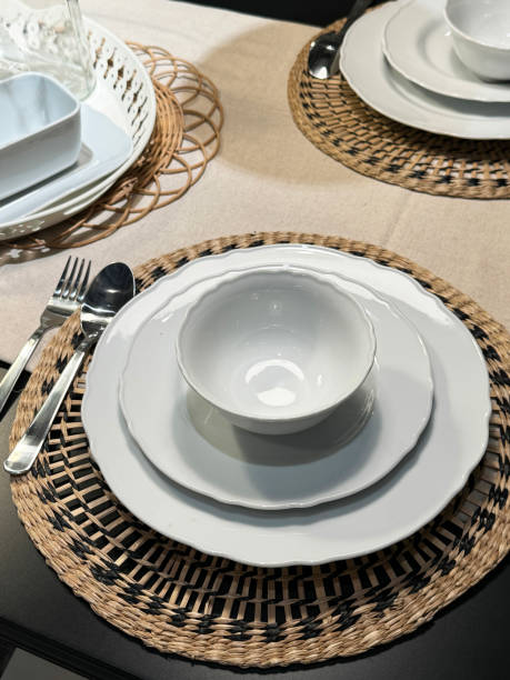 istock Close-up image of dining table set for breakfast / lunch / dinner with stacked white dinner plates, side plates and bowls on round wicker placemats, stainless steel cutlery spoons and forks, elevated view, focus on foreground 2148415097