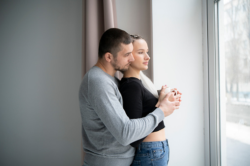 Man and Woman Standing by Window looking outside, drinking hot tea together