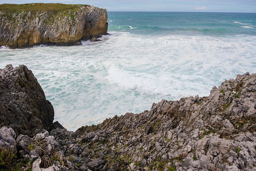 A rocky cliff juts out over the water, with sharp rocks and jagged edges. The body of water in the background stretches out to the horizon under a clear sky.