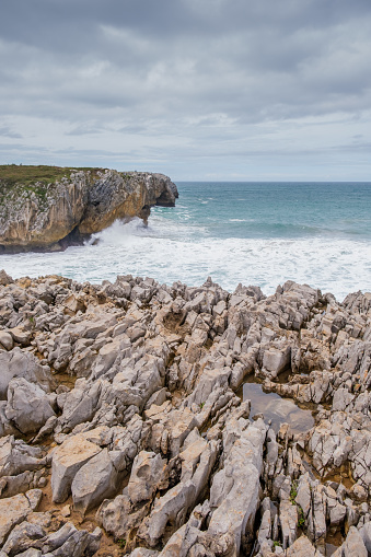 A view of a rocky beach with sharp rocks jutting out of the water in Los Bufones de Llanes, Asturias. The waves crash against the rugged coastline, creating a dynamic and powerful scene.