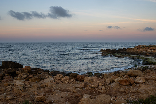 Calm dark blue water of Mediterranean sea. Curve of the rocky part of the coast. Orange and blue sky at the sunrise. Darker blue clouds. Spiaggia Sibiliana, Marsala, Sicily, Italy.