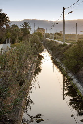 Ruin of a building Casa dell Acqua. Channel from it to the sea. Reflection of an early morning sky. Streetlamps lining the canal, together with road. Petrosino, Sicily, Italy.