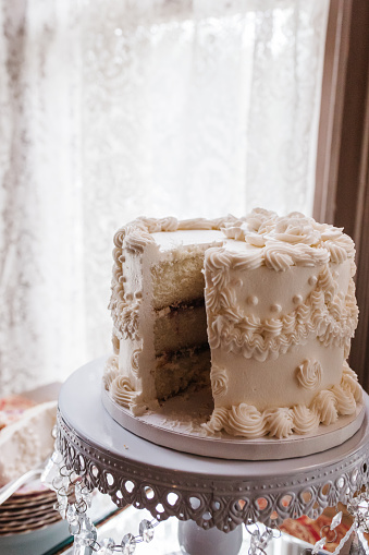 This image presents a wedding cake with a classic touch, set against a delicate lace curtain and soft natural lighting. The cake features traditional white icing with intricate piping designs, showcasing a partially sliced view that reveals its layered interior.