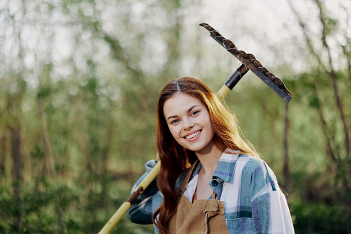 A woman smiling beautifully and looking at the camera, a farmer in work clothes and an apron working outdoors in nature and holding a rake to gather grass and forage for the animals in the garden. High quality photo