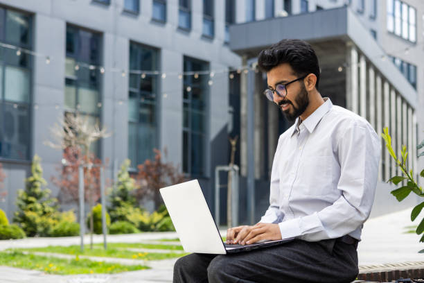 istock Smiling young businessman using laptop outdoors with modern building backdrop 2148421376