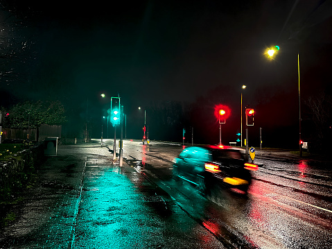 Pontypridd, Wales - 14 March 2024: Set of traffic lights at a road junction on a wet night with motion blur of  a car driving at speed
