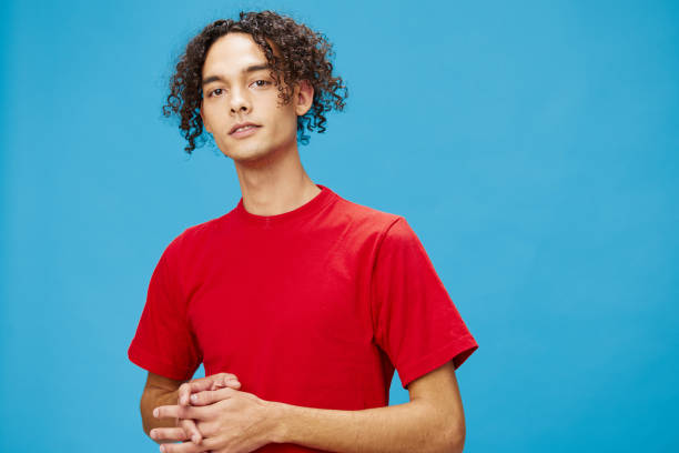 istock Attractive cute young curly tanned Caucasian guy in basic red t-shirt looks at camera posing isolated on over blue background. Lifestyle and Emotions concept. Good offer with copy space for ad 2148426092