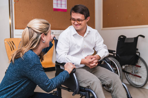 Empathetic Encounter: Woman Connects with Man in Wheelchair with Down Syndrome A heartfelt chat between a woman and a smiling man with Down Syndrome in a wheelchair. physical disability stock pictures, royalty-free photos & images