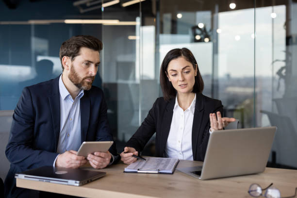 Successful business partners sitting by table with digital devices and looking at pc screen attentively. Caucasian woman and man in formalwear discussing and analyzing statistics of sale season