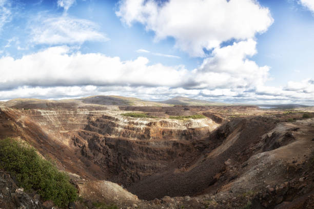 A rugged rock outcrop and narrow ridge dominate the foreground, with jagged, layered stone and sparse shrubs clinging to crevices. The viewpoint overlooks densely forested rolling hills and distant mountains under a bright summer sky, creating a panoramic, tranquil natural landscape.