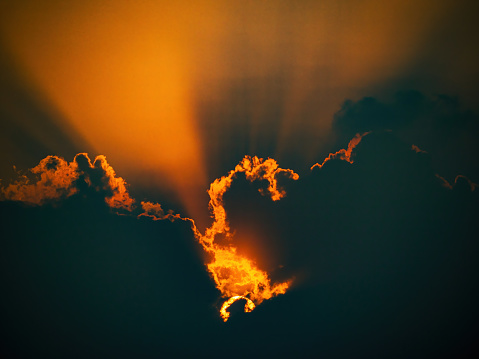 The sun is setting behind a cloud. The sky is orange and the clouds are dark. The sun is shining through the clouds, creating a beautiful and serene scene