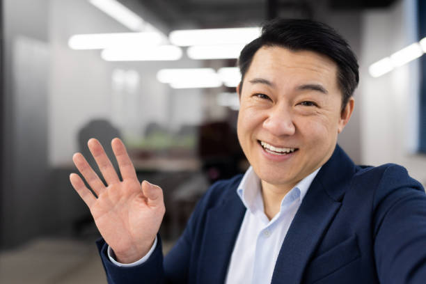 istock Friendly japanese employer in strict outfit waving hello while looking at camera with toothy smile. Positive entrepreneur connecting to group video meeting by digital gadget with newly hired trainees 2148432969