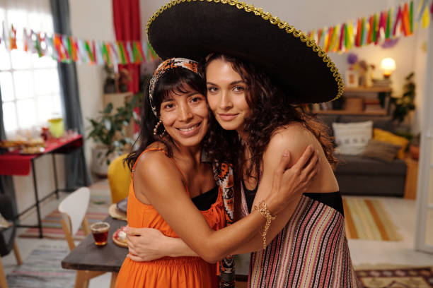 Two happy young intercultural girlfriends in national attire embracing and looking at you while standing in front of camera in living room