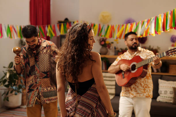 Rear view of young dynamic woman with dark long wavy hair dancing in front of two Hispanic men playing musical instruments at home party
