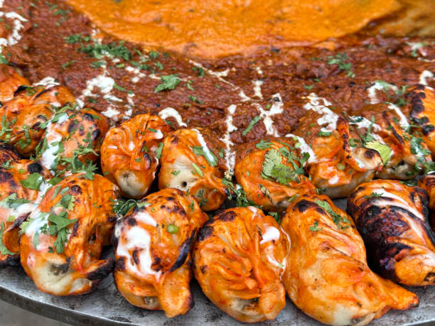 Stock photo showing close-up, elevated view of popular Indian street food tikka momos (South Asian dumplings) served with tandoori and tikka curry gravy sold by street food vendors on streets or market stalls in India. imagen de fotograma completo de tikka momos (albóndigas del sur de asia) servidas con salsa de curry tandoori y tikka, comida callejera preparada en tawa (sartén grande y plana de metal), puesto de comida salada tradicional india, vista elevada, enfoque