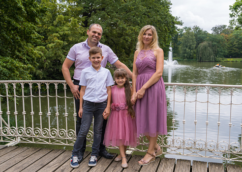 Portrait of a happy family. Laughing dad and mom, teenage son and first-grader daughter stand on a white bridge in the park. Mom and girl are dressed in beautiful pink dresses