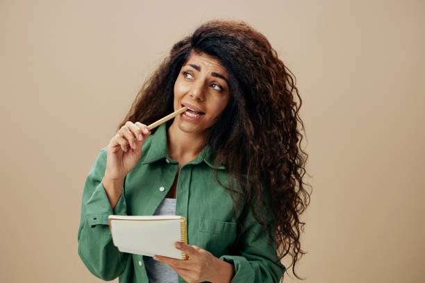 istock Stressed thoughtful pensive tanned curly Latin female in casual things with notebook chew pencil posing isolated over pastel beige background look up. Copy space Mockup Banner. Good offer 2148438043