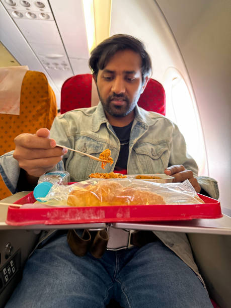 istock Close-up image of Indian man in plane seat with fold down table containing plastic tray of chicken kebab sausage with fried rice and roti (traditional Indian flatbread), plastic bottle of water, wooden fork cutlery, focus on foreground 2148438240