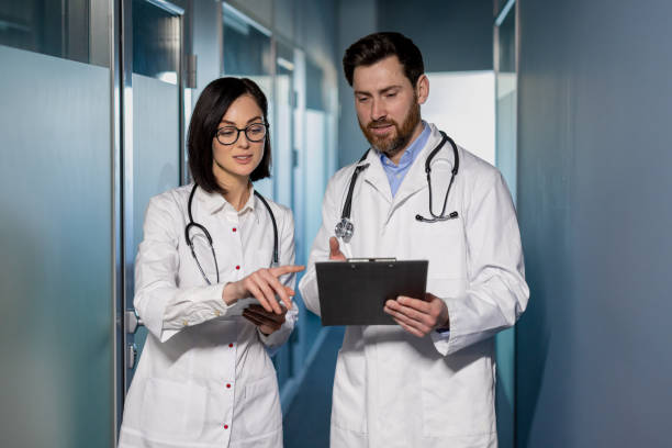 istock Two professional doctors in white lab coats looking at clipboard in hands while standing in hall of clinic. Coworkers discussing medical history of patient and forming new treatment strategy 2148438325
