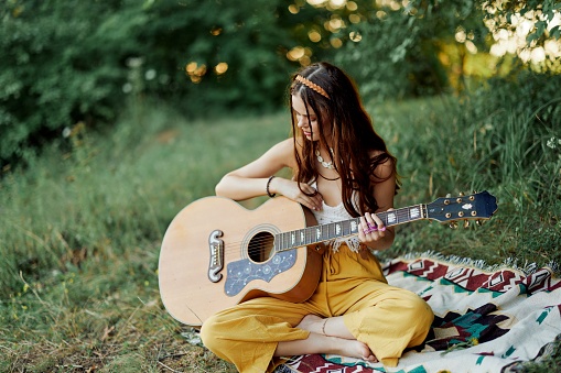 Girl hippie woman playing guitar in eco-friendly clothes sitting on the ground outside in nature in the fall watching the sunset. High quality photo