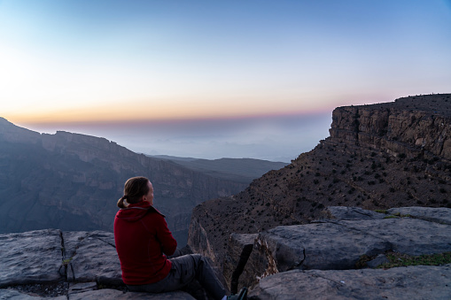 One women, rear view, watching sunset, sitting at the edge at the edge of Oman - Grand Canyon/Wadi Nakha. In distance some unrecognizable persons.