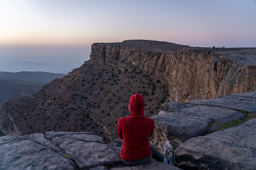 One women, rear view, watching sunset, sitting at the edge at the edge of Oman - Grand Canyon/Wadi Nakha. In distance some unrecognizable persons.