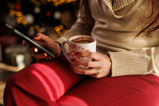 Midsection of unrecognizable young woman sitting by the window, in front of the glistening Christmas tree, enjoying a cup of coffee, scrolling online via smart phone.