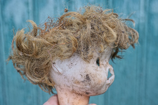 hand holds the head of an old brown broken plastic doll with dirty hair on a green background