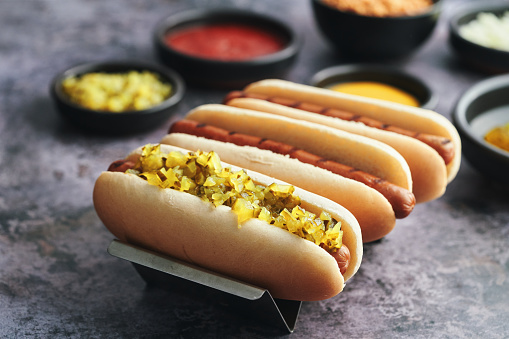 The photo depicts a mouthwatering assortment of Hot Dogs, each one customized with roasted onions, pickle relish and a variety of toppings.