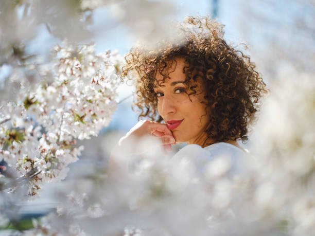 A 9-year-old girl with curly hair poses next to a bouquet of flowers, looking at the camera, in a light-filled, white-toned setting.