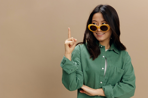 Hey, good purpose. Smiling Korean young woman in khaki green shirt stylish eyewear point finger up posing isolated on over beige pastel studio background. Cool fashion offer. Sunglasses ad concept