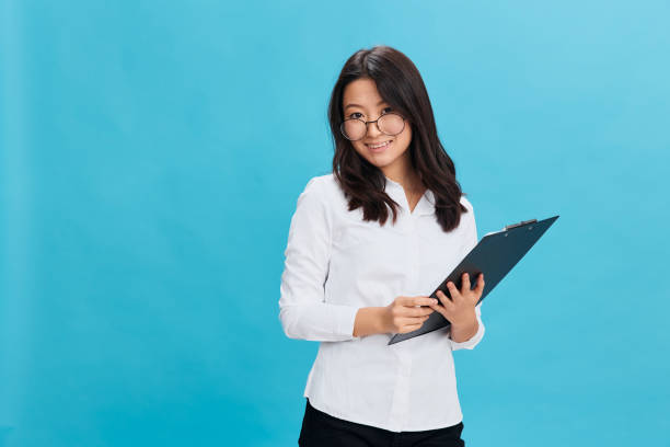 istock Friendly cute Asian businesswoman in classic office dress code holds folder tablet with applicant questionnaire posing isolated on blue studio background. Cool business offer. Great career concept 2148443495