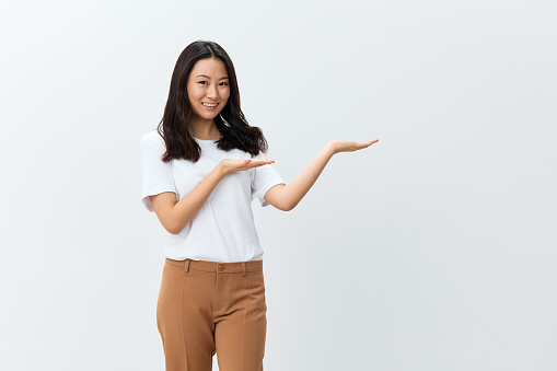 Hey, look here. Attractive Asian Korean young female in white basic t-shirt hold palms up show free place posing isolated on white studio background. The best offer for ad. People Lifestyle concept