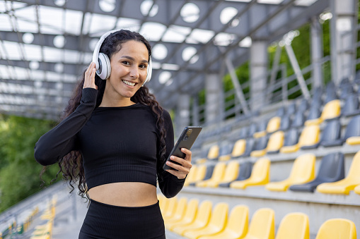 A smiling hispanic woman wearing headphones and sportswear, holding a smartphone, enjoying music in a sunny stadium setting.