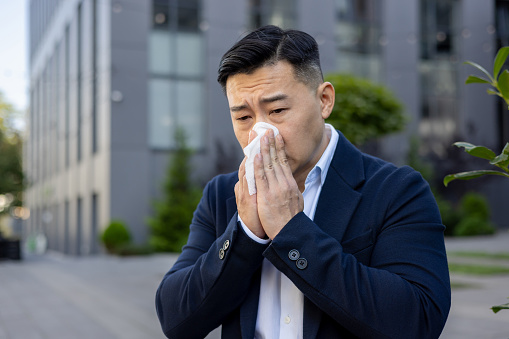 Close-up photo of a sick young Asian male businessman sitting on a bench near an office building and wiping his nose, with a napkin from a runny nose.
