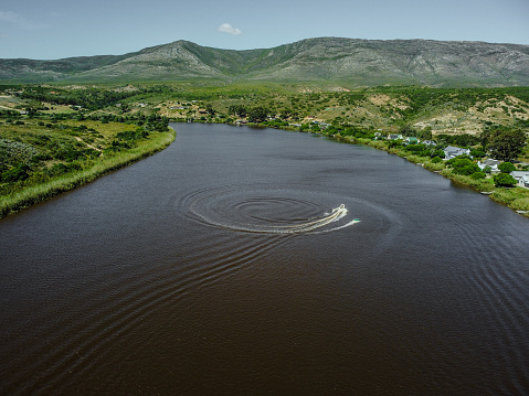 Aerial shot of a pontoon boat moving around in circles towing a recreational tube with multiple people on both the tube and the boat on the brown Breerivier surface, with a view of multiple waterfront houses on the lush green river banks and a mountain range in the background, Malgas.